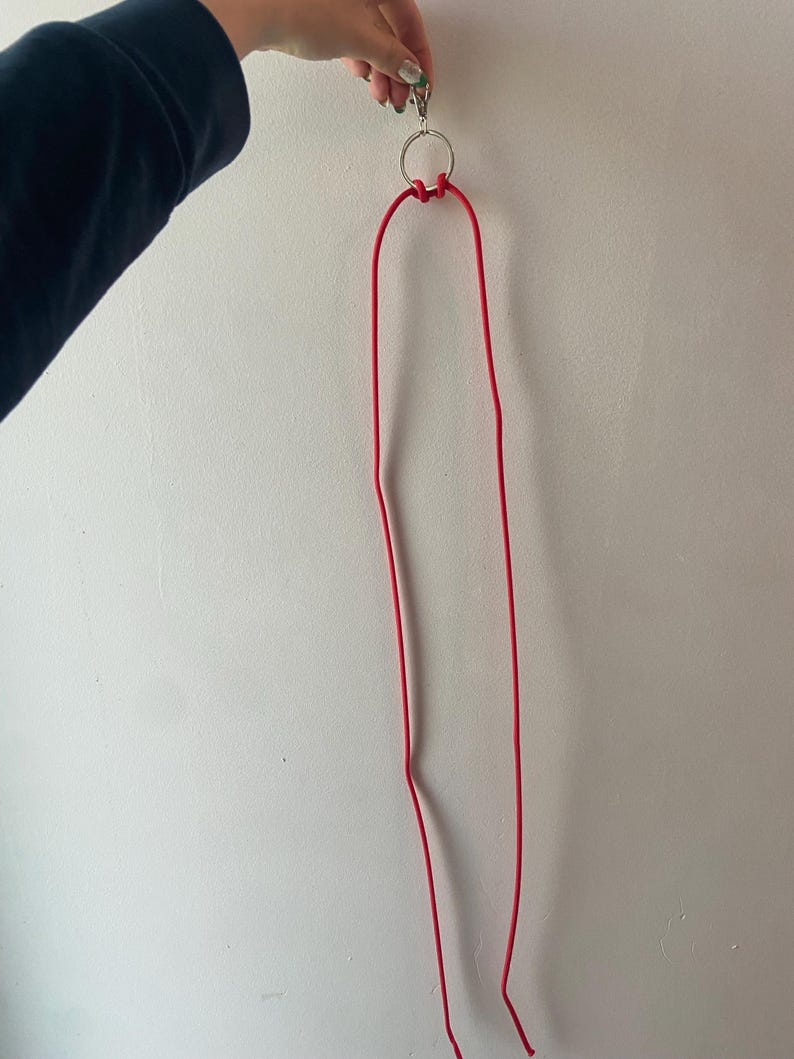 May include: A red lanyard with a silver metal ring and clasp. The lanyard is made of a thin, flexible material and is hanging against a white wall. The lanyard is held by a hand.