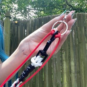 May include: A red and white braided leash with a black clasp and a silver keyring. The leash is held up against a wooden fence and green foliage.