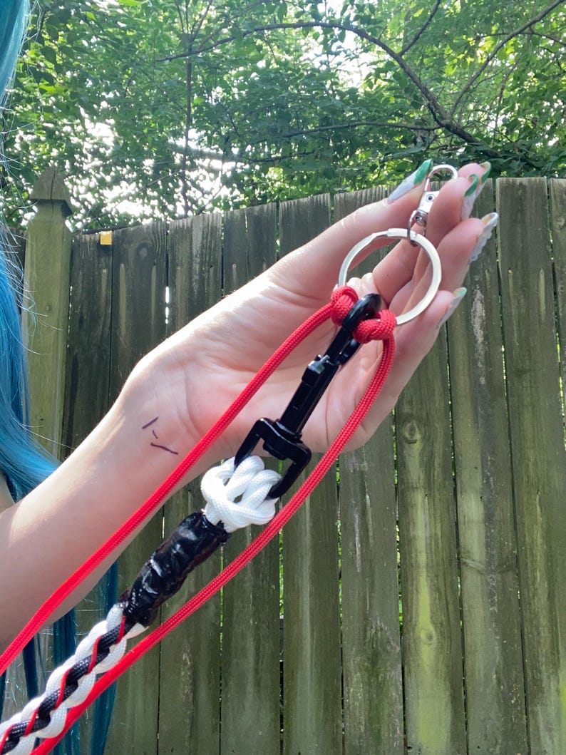 May include: A red and white braided leash with a black clasp and a silver keyring. The leash is held up against a wooden fence and green foliage.