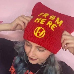 Könnte beinhalten: Eine rote Häkelmütze mit dem gelben Schriftzug "FROM HERE" und einem gelben Symbol. Die Mütze wird von einer Person hochgehalten. Der Hintergrund ist einfarbig rosa.