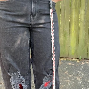 May include: Black denim jeans with distressed details, featuring ripped knees and a decorative braided strap with red and white accents. The jeans have a classic five-pocket design and a black belt.