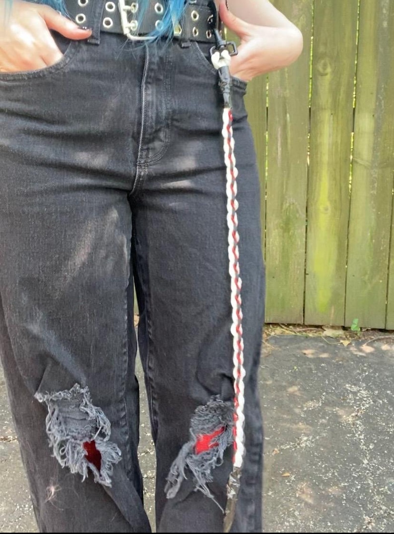 May include: Black denim jeans with distressed details, featuring ripped knees and a decorative braided strap with red and white accents. The jeans have a classic five-pocket design and a black belt.