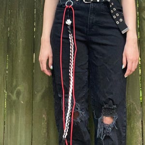 May include: A black belt with silver grommets and a silver buckle. Attached is a red and white lanyard with a decorative charm and a braided white and black cord. The lanyard hangs down the front of black ripped jeans.