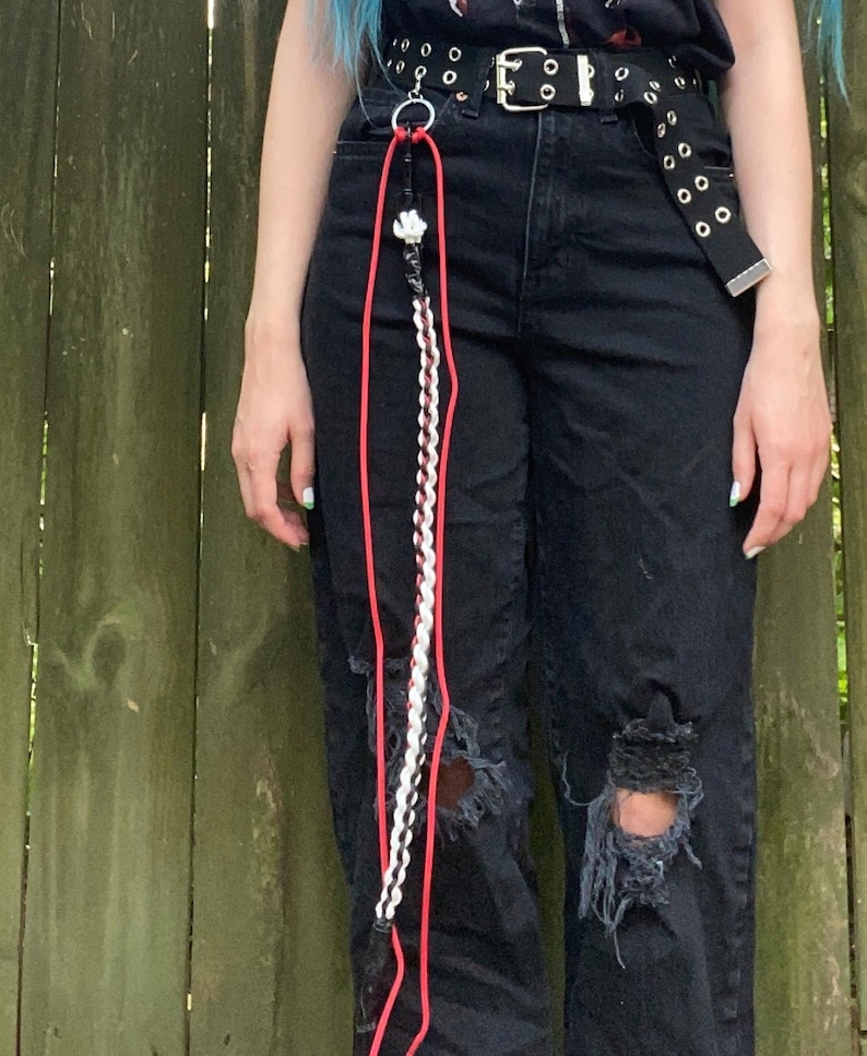 May include: A black belt with silver grommets and a silver buckle. Attached is a red and white lanyard with a decorative charm and a braided white and black cord. The lanyard hangs down the front of black ripped jeans.