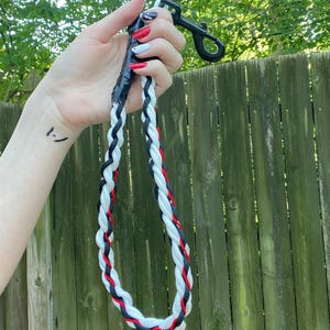 May include: A braided dog leash in black, white, and red. The leash is held by a black clip and is made of interwoven rope. The leash is approximately 48 inches long. The background is a wooden fence and green foliage.
