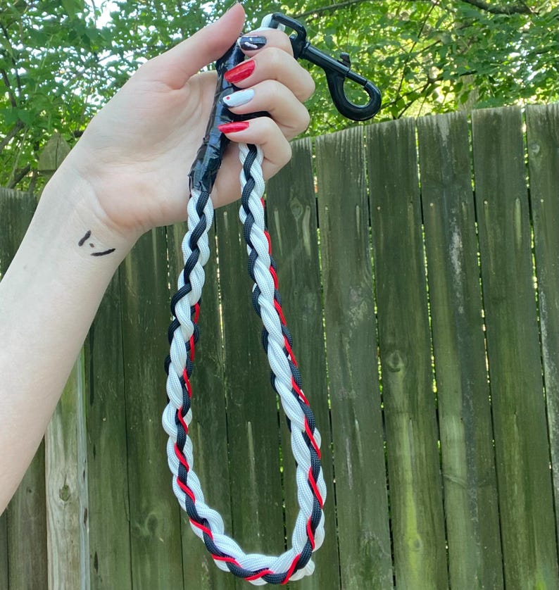 May include: A braided dog leash in black, white, and red. The leash is held by a black clip and is made of interwoven rope. The leash is approximately 48 inches long. The background is a wooden fence and green foliage.