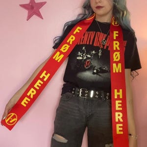 May include: A red sash with the words "FROM HERE" in yellow, featuring a band logo. The sash is worn over a black t-shirt with a band graphic and black jeans. A pink star is visible in the background.