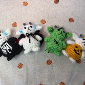 May include: Four small, handmade crochet plush toys featuring goat-like creatures. One wears a black skeleton suit, one a black cape, one is green with stitched details, and one wears a yellow pumpkin costume. Each has small horns and blue accents.