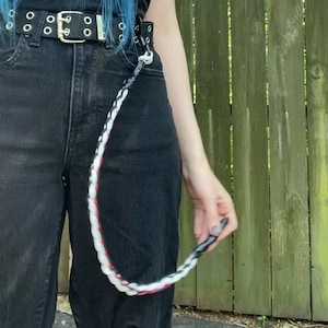 May include: A black belt with silver grommets and a matching black and white braided chain. The chain has red accents and is attached to the belt loop. The person is wearing black jeans.