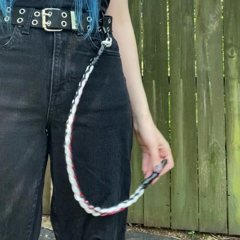 May include: A black belt with silver grommets and a matching black and white braided chain. The chain has red accents and is attached to the belt loop. The person is wearing black jeans.
