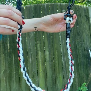May include: Braided leash featuring black, white, and red strands. The leash has a black clip and a black handle. The leash is held up against a wooden fence.