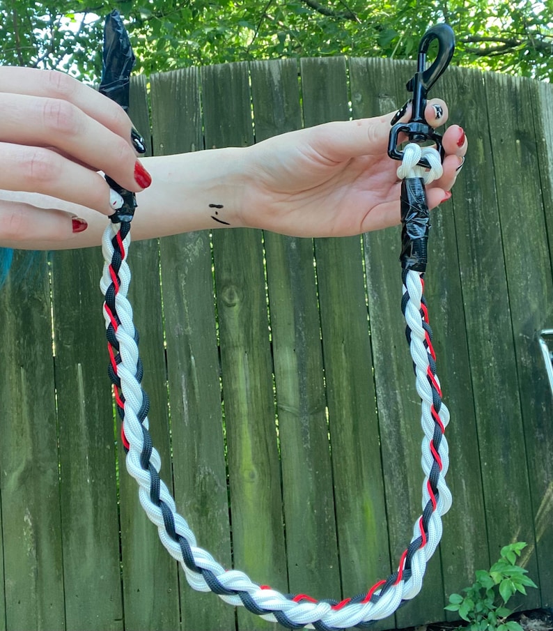 May include: Braided leash featuring black, white, and red strands. The leash has a black clip and a black handle. The leash is held up against a wooden fence.