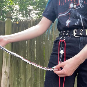 May include: A braided leash in black, white, and red is held by a person wearing a black t-shirt and black jeans. The leash is attached to a red lanyard with a black clip and a white charm. The person is standing in front of a wooden fence.