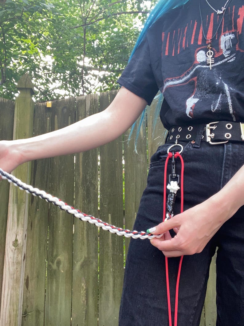 May include: A braided leash in black, white, and red is held by a person wearing a black t-shirt and black jeans. The leash is attached to a red lanyard with a black clip and a white charm. The person is standing in front of a wooden fence.