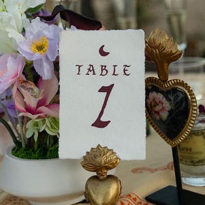 May include: A wedding table setting with a floral arrangement in a white vase, a table number card, and decorative heart-shaped objects. The table number card reads "Table 1". The setting is on a patterned tablecloth.