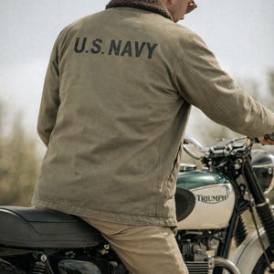 Puede incluir: Chaqueta verde oliva con "U.S. NAVY" impresa en la espalda. La chaqueta tiene un cuello de piel marrón. La persona está sentada en una motocicleta Triumph con un depósito verde y blanco.