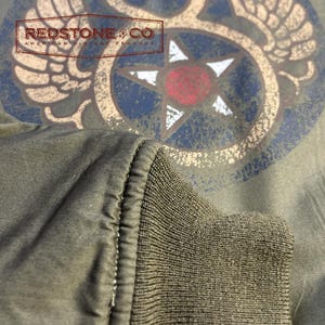 May include: Close-up of an olive green jacket sleeve with a ribbed cuff. The jacket features a distressed graphic design with a star and wings, and the text "REDSTONE & CO AMERICAN VINTAGE PRODUCT".