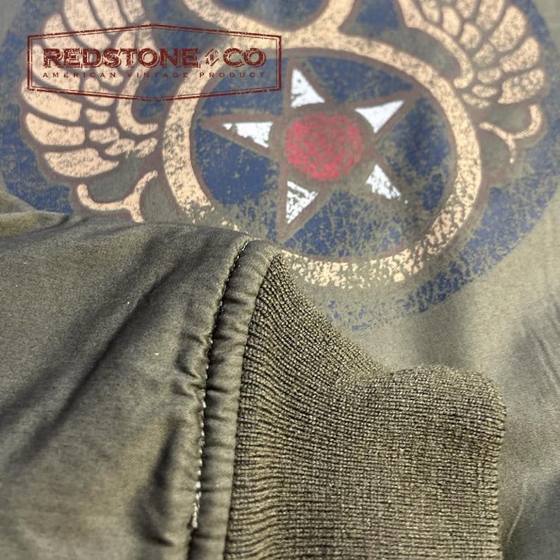 May include: Close-up of an olive green jacket sleeve with a ribbed cuff. The jacket features a distressed graphic design with a star and wings, and the text "REDSTONE & CO AMERICAN VINTAGE PRODUCT".
