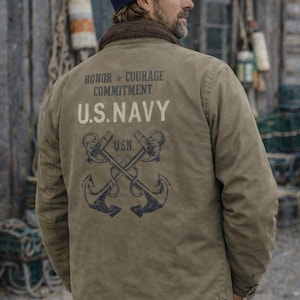 Puede incluir: Un hombre con gorro azul marino y chaqueta verde caqui. La chaqueta tiene la inscripción "HONOR + COURAGE COMMITMENT U.S. NAVY" en la espalda, con anclas cruzadas y las letras "U.S.N."