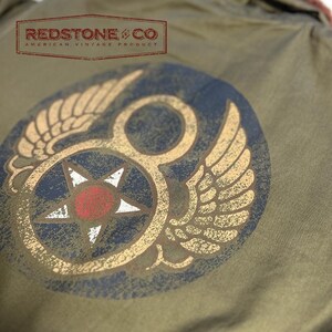 May include: Olive green garment with a distressed emblem featuring wings, a star, and the text "REDSTONE & CO AMERICAN VINTAGE PRODUCT". The emblem has gold, red, white, and navy blue accents. The garment has a vintage aesthetic.