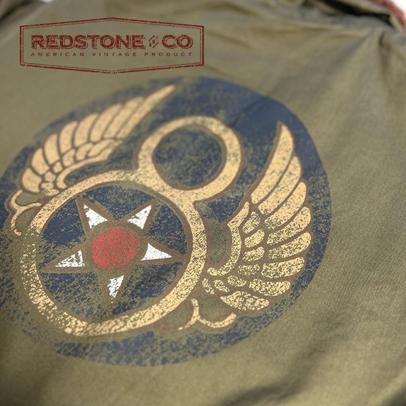 May include: Olive green garment with a distressed emblem featuring wings, a star, and the text "REDSTONE & CO AMERICAN VINTAGE PRODUCT". The emblem has gold, red, white, and navy blue accents. The garment has a vintage aesthetic.