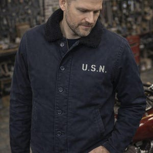 Op de afbeelding: Marineblauwe jas met een zwarte imitatiebontkraag en knoopsluiting. De jas heeft de tekst "U.S.N." in het wit op de borst. De jas wordt gedragen met een donkerblauwe jeans.