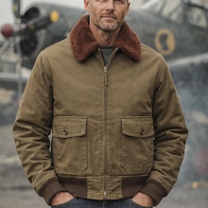 May include: An olive green bomber jacket with a brown faux fur collar, two front pockets, and ribbed cuffs and hem. The jacket is zipped up and worn with blue jeans. An airplane is in the background.
