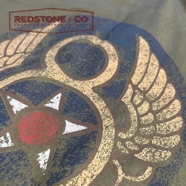 May include: Close-up of an olive green t-shirt with a distressed design. It features a compass rose, wings, and the text "REDSTONE & CO AMERICAN VINTAGE PRODUCT". The design incorporates red, white, and tan colours.