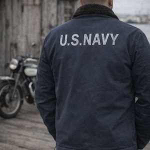 Pode incluir: Casaco azul marinho com o texto "U.S. NAVY" em branco nas costas. O casaco tem uma gola castanha escura e é usado com jeans azuis. Uma motocicleta vintage está estacionada perto.