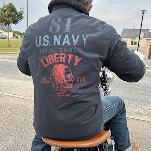 Puede incluir: Una chaqueta negra con un parche azul marino que dice "81 U.S. NAVY U.S.S. SABLE PATROL LIBERTY" con un gráfico rojo y blanco de una cabeza de nativo americano y el texto "USA" debajo.