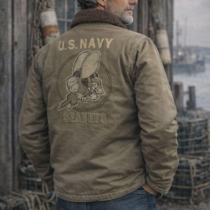 Pode incluir: Casaco verde-oliva com gola de lã castanha. A parte de trás do casaco apresenta o texto "U.S. NAVY" acima de um desenho animado de uma abelha segurando uma chave, com a palavra "SEABEES" abaixo. A pessoa está usando jeans azuis.