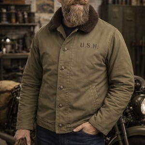 Puede incluir: Chaqueta verde oliva con cuello de piel marrón y letras "U.S.N." en el pecho. La chaqueta abotonada se combina con vaqueros azules. La persona sostiene guantes marrones. El fondo sugiere un taller.
