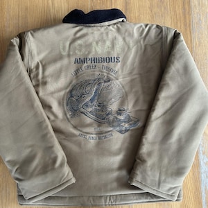 Peut inclure: Une veste USMC Amphibious marron avec un col noir et un graphique d'un alligator et d'un char. Le texte sur la veste est "USMC Amphibious Little Creek Virginia 62nd Naval Beach Battalion".