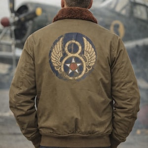 May include: An olive green bomber jacket with a brown fur collar. The back of the jacket displays a circular emblem with wings, the number 8, and a star. The person is wearing blue jeans. An aeroplane is in the background.