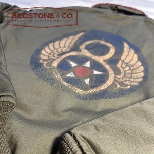 May include: Olive green bomber jacket with a distressed vintage appearance. It has a large emblem on the back with wings, a star, and the number 8. The text "REDSTONE & CO AMERICAN VINTAGE PRODUCT" is printed at the top.