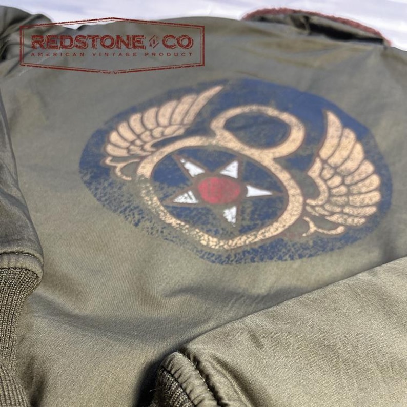 May include: Olive green bomber jacket with a distressed vintage appearance. It has a large emblem on the back with wings, a star, and the number 8. The text "REDSTONE & CO AMERICAN VINTAGE PRODUCT" is printed at the top.