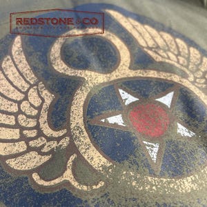 May include: Close-up of a distressed olive green fabric with a vintage-style graphic. The design features a central star with a red centre, surrounded by a blue circle and tan wings. The text "REDSTONE & CO AMERICAN VINTAGE PRODUCT" is at the top.