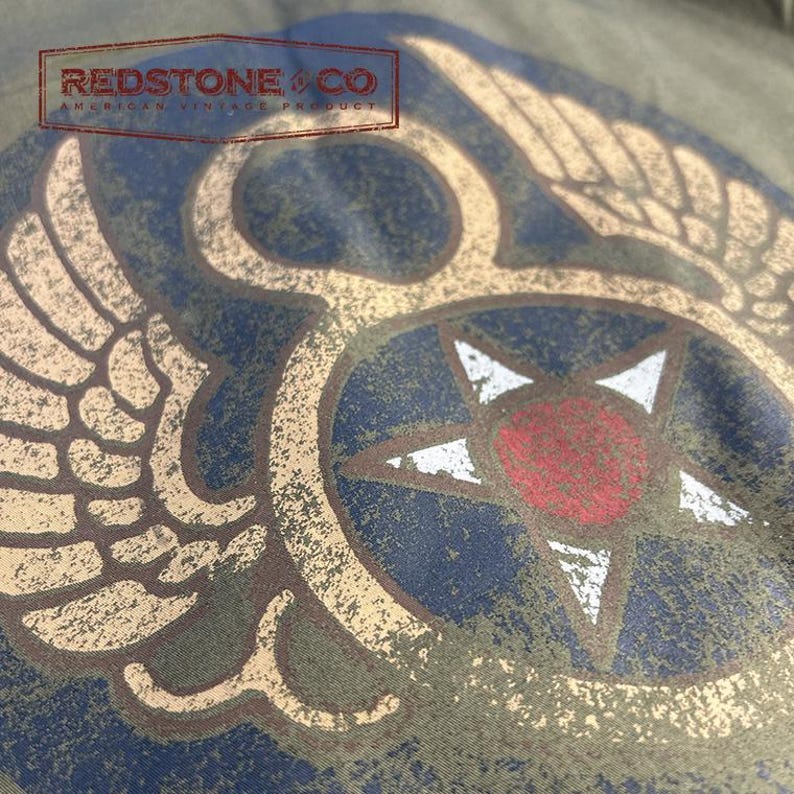 May include: Close-up of a distressed olive green fabric with a vintage-style graphic. The design features a central star with a red centre, surrounded by a blue circle and tan wings. The text "REDSTONE & CO AMERICAN VINTAGE PRODUCT" is at the top.