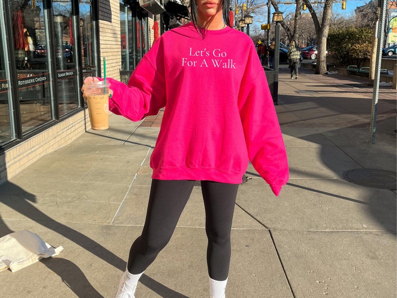 May include: A woman wearing a pink sweatshirt with the text "Let's Go For A Walk" in white. She is holding a to-go cup of coffee.