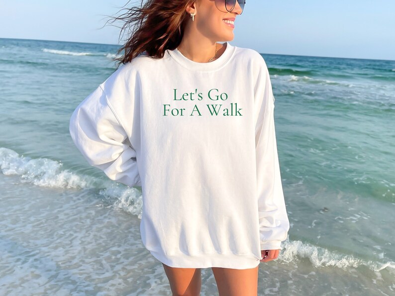 May include: White sweatshirt with the text "Let's Go For A Walk" in green lettering. The sweatshirt is worn by a person standing on a beach.