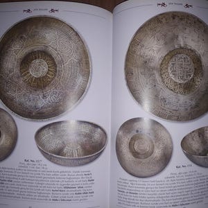 May include: Two open book pages displaying antique metal bowls and plates. The bowls and plates have intricate engraved designs. The book pages also contain text in a foreign language. The items are silver in color.