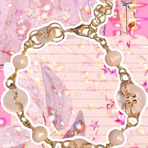 May include: A gold-toned chain bracelet featuring large, pearlescent beads and button-shaped charms. The bracelet is set against a pink background with a collage of paper and glitter effects, creating a whimsical aesthetic.