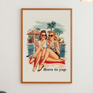May include: A framed vintage style poster featuring two women in sunglasses by a pool, holding drinks. They are sitting on a red towel. Palm trees and a cabana are in the background. The text 'Born to yap' is printed below the image. The poster has a light beige background and a wooden frame.