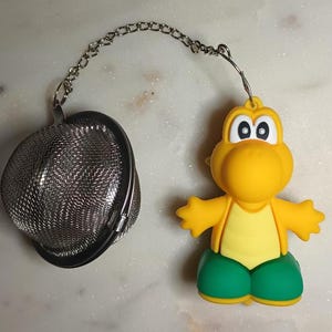 May include: A novelty tea infuser featuring a yellow cartoon character with green shorts. The infuser has a stainless steel mesh basket attached to a chain. The character is a Koopa Troopa from the Super Mario Bros. video game series.