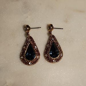 May include: A pair of gold-toned drop earrings. Each earring features a teardrop-shaped, dark blue gemstone surrounded by small, clear crystals and an ornate gold filigree design. The earrings have post-style closures.