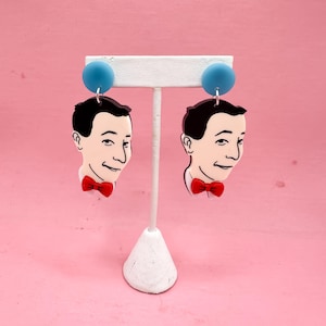 Könnte beinhalten: Ein Paar ausgefallene Ohrringe mit einem Cartoon-Gesicht. Jeder Ohrring hat ein schwarzhaariges Gesicht mit roter Fliege und einer blauen Kugel oben. Auf einem weißen Ständer vor rosa Hintergrund.