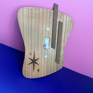May include: A wooden light switch cover with a mid-century modern design. The cover is light brown with a starburst design and two vertical brown accent pieces. The switch is white, and the cover is mounted on a pink and blue wall.