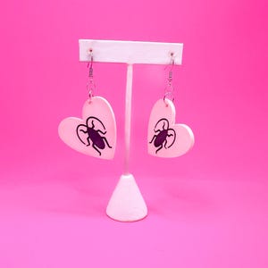 May include: A pair of heart-shaped earrings with a black insect design on a light pink background. The earrings are displayed on a white earring stand against a bright pink backdrop. The earrings have silver-colored hooks.