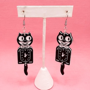 Pode incluir: Um par de brincos em forma de gato em preto e branco. Cada brinco apresenta a cabeça de um gato acima de um mostrador de relógio, com uma pequena gravata borboleta e uma cauda. Os brincos são suspensos por ganchos prateados sobre um fundo rosa.