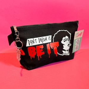 Puede incluir: Bolsa de cosméticos negra con el texto "DON'T DREAM IT BE IT" en letras blancas y rojas goteando. Un gráfico en blanco y negro de una persona fumando está a la derecha. Un llavero con un dije de labio rojo cuelga de la cremallera.
